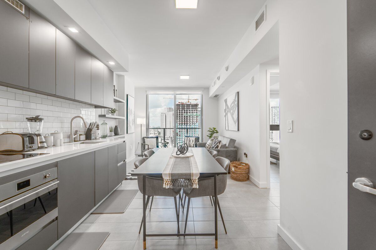 A modern kitchen with grey cabinets, a dining table, and a view of a cityscape through large windows.