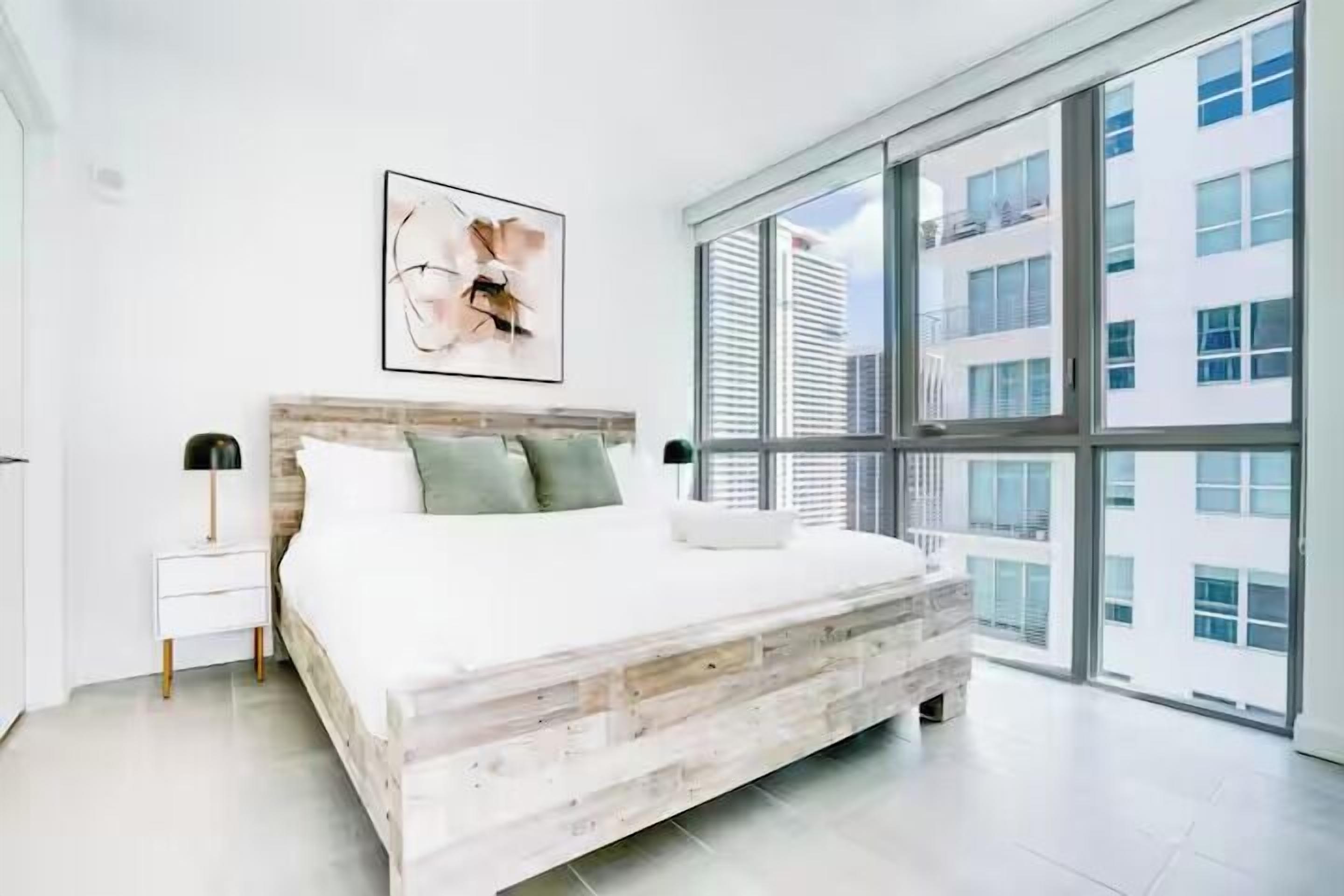 Un dormitorio moderno con una cama grande, una mesita de noche, una lámpara de pared y vistas a la ciudad a través de ventanales de gran altura.