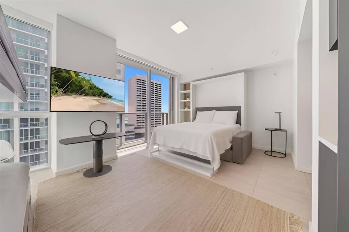 A modern bedroom with a large bed, a TV, and a balcony with a city view.