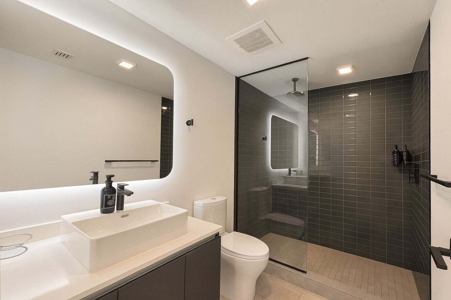Un baño moderno con un espejo grande, un lavamanos blanco, un inodoro y una zona de ducha con mampara de vidrio.