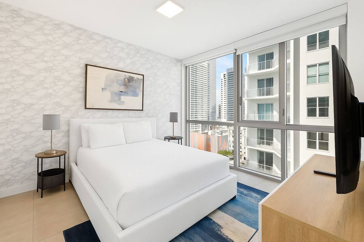 A modern hotel room with a large bed, two bedside tables, a TV, and a large window offering a city view.
