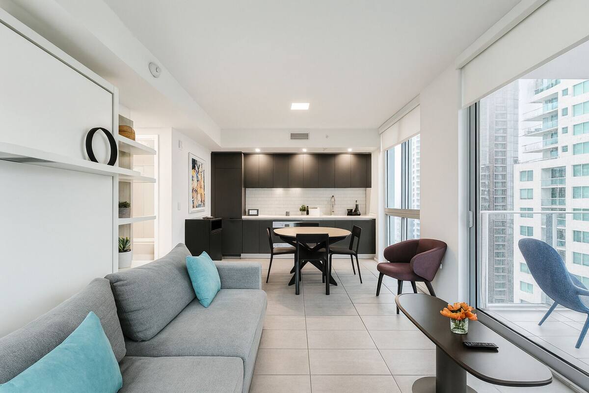 A modern apartment with a grey sofa, a dining table, and a view of a cityscape through large windows.