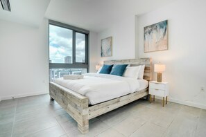 A modern bedroom with a large bed, a nightstand, a window with a city view, and a painting on the wall. - Miami Downtown Condos (Miami)