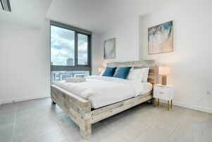 A modern bedroom with a large bed, a nightstand, a window with a city view, and a painting on the wall.