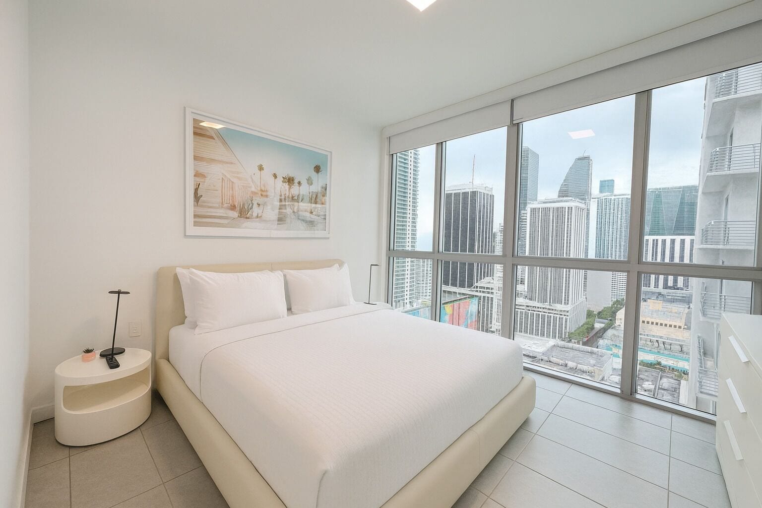 A modern bedroom with a large bed, a bedside table with a lamp, a cityscape view through large windows, and a framed picture on the wall.