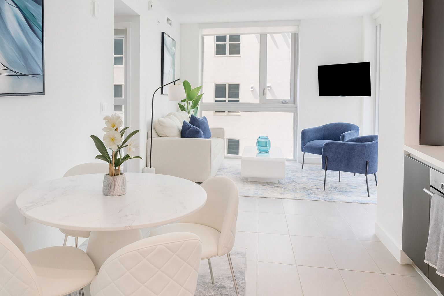 Un salon moderne avec un canapé blanc, des fauteuils bleus, une table ronde avec des chaises, une table basse et un téléviseur fixé au mur.