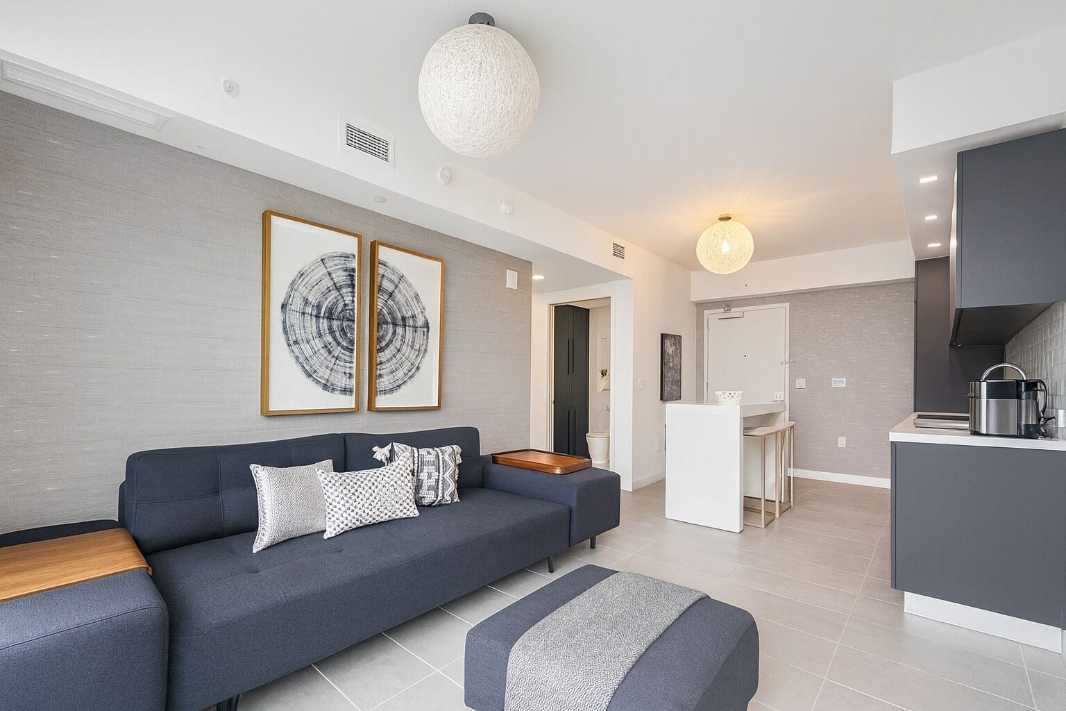 A modern living room with a dark blue sofa, a white coffee table, and a grey ottoman. There are two framed artworks on the wall, a kitchen area with a white countertop and a grey cabinet, and a large round light fixture on the ceiling.