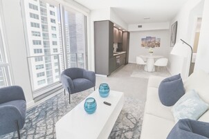 A modern living room with a sofa, armchairs, and a coffee table. There is a dining area with a table and chairs, and a kitchen in the background.