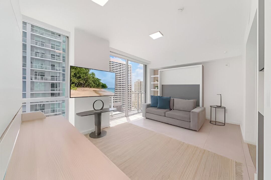 A modern living room with a large TV, a grey sofa, and a view of tall buildings.