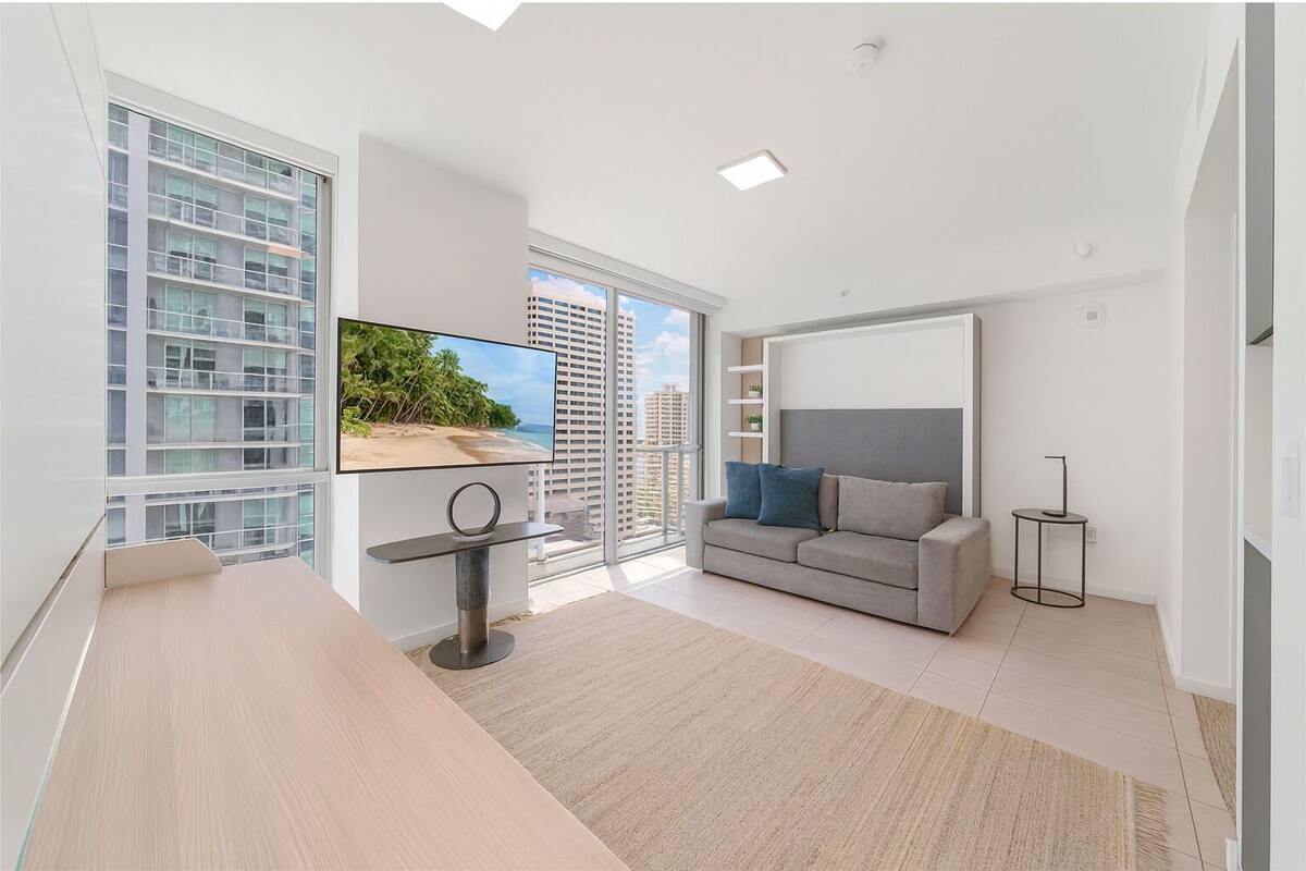 A modern living room with a large TV, a grey sofa, and a view of a beach and buildings through large windows.