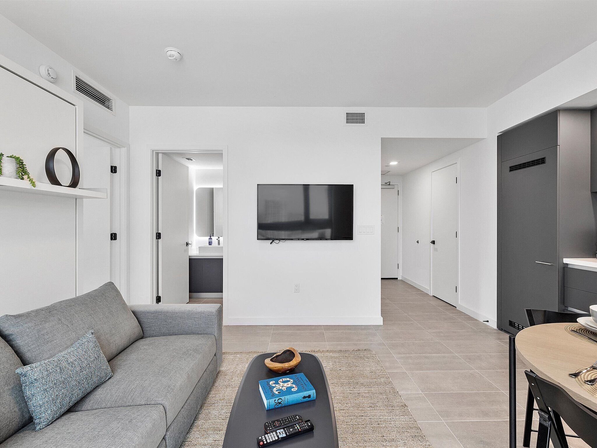 A modern living room with a grey sofa, a coffee table with a book and remote, a TV on the wall, and a kitchen area visible through an open door.
