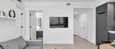 A modern living room with a grey sofa, a coffee table with a book and remote, a TV on the wall, and a kitchen area visible through an open door.