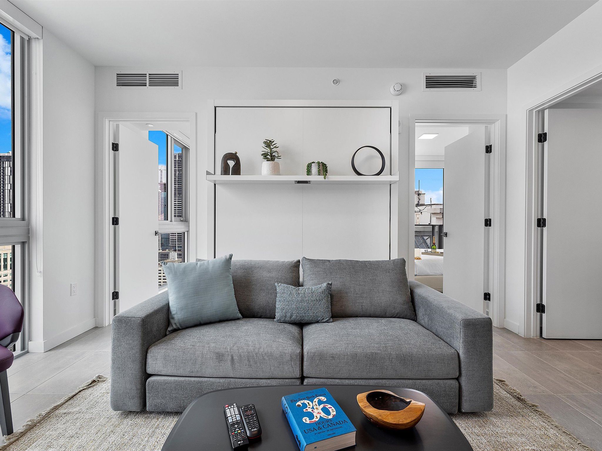 Un salon moderne avec un canapé d’angle gris, une table basse sur laquelle se trouvent des télécommandes et un livre, et un mur blanc avec une étagère au-dessus.