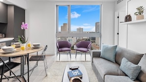 Ein modernes Wohnzimmer mit einem grauen Sofa, zwei violetten Sesseln, einem Esstisch mit Tellern und Gläsern sowie einem großen Fenster mit Blick auf die Stadt.