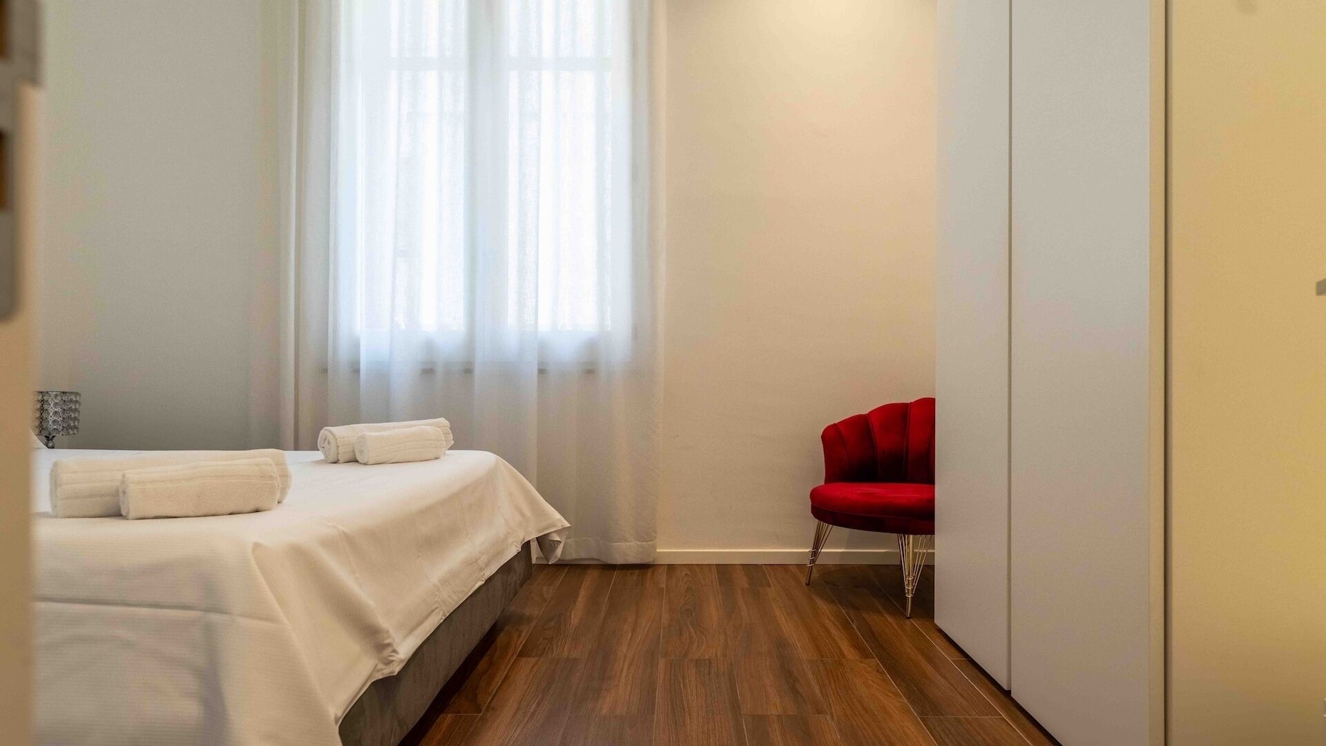 A hotel room with a bed, a red chair, and a window with sheer curtains.