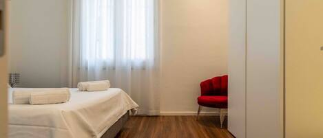 A hotel room with a bed, a red chair, and a window with sheer curtains.