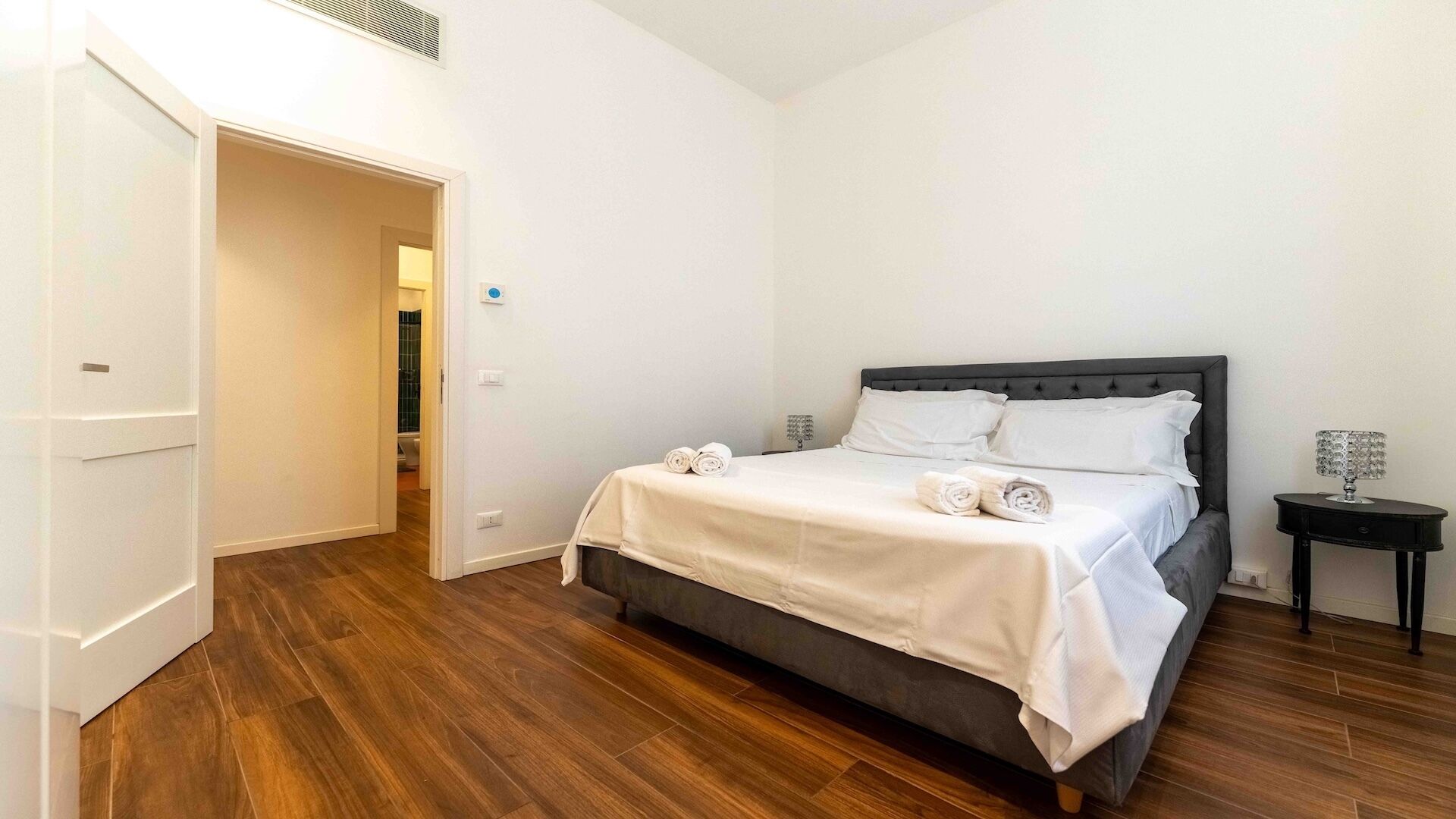 A hotel room with a bed, a bedside table, a lamp, and a door leading to another room.