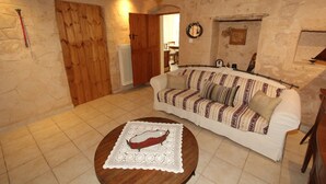 A cozy living room with a wooden floor, a white sofa with patterned cushions, a wooden coffee table with a lace table runner, and a stone wall with a wooden door. - Kipseli s Stone House by Frangki (Aegina)