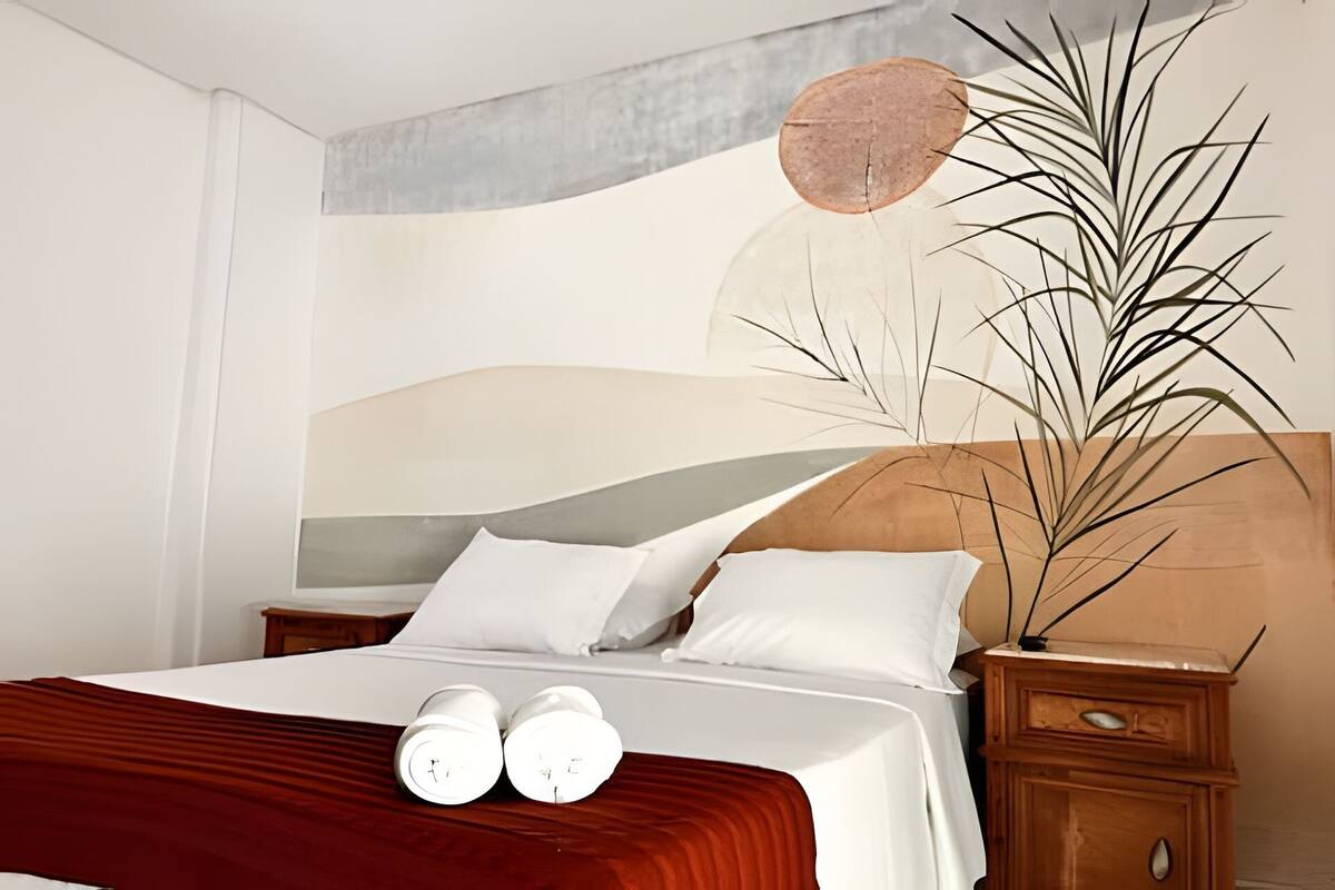Uma cama bem arrumada com lençóis brancos, um cobertor vermelho e duas garrafas brancas ao lado.