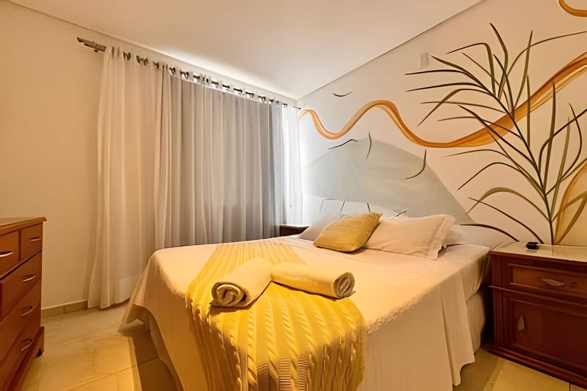 Quarto de hotel com cama, cómoda de madeira, mesa de cabeceira e parede com planta e arte abstrata.