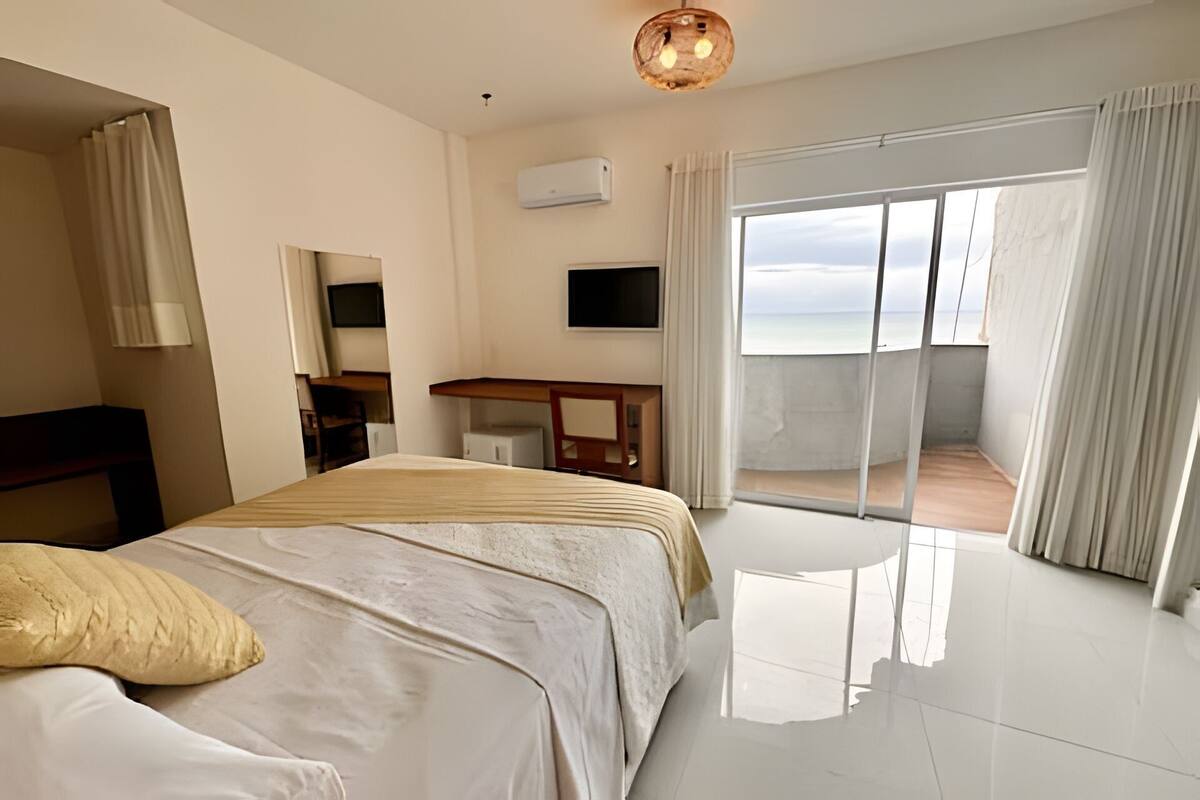 Quarto de hotel com cama, escrivaninha, televisão e varanda com vista para o mar.