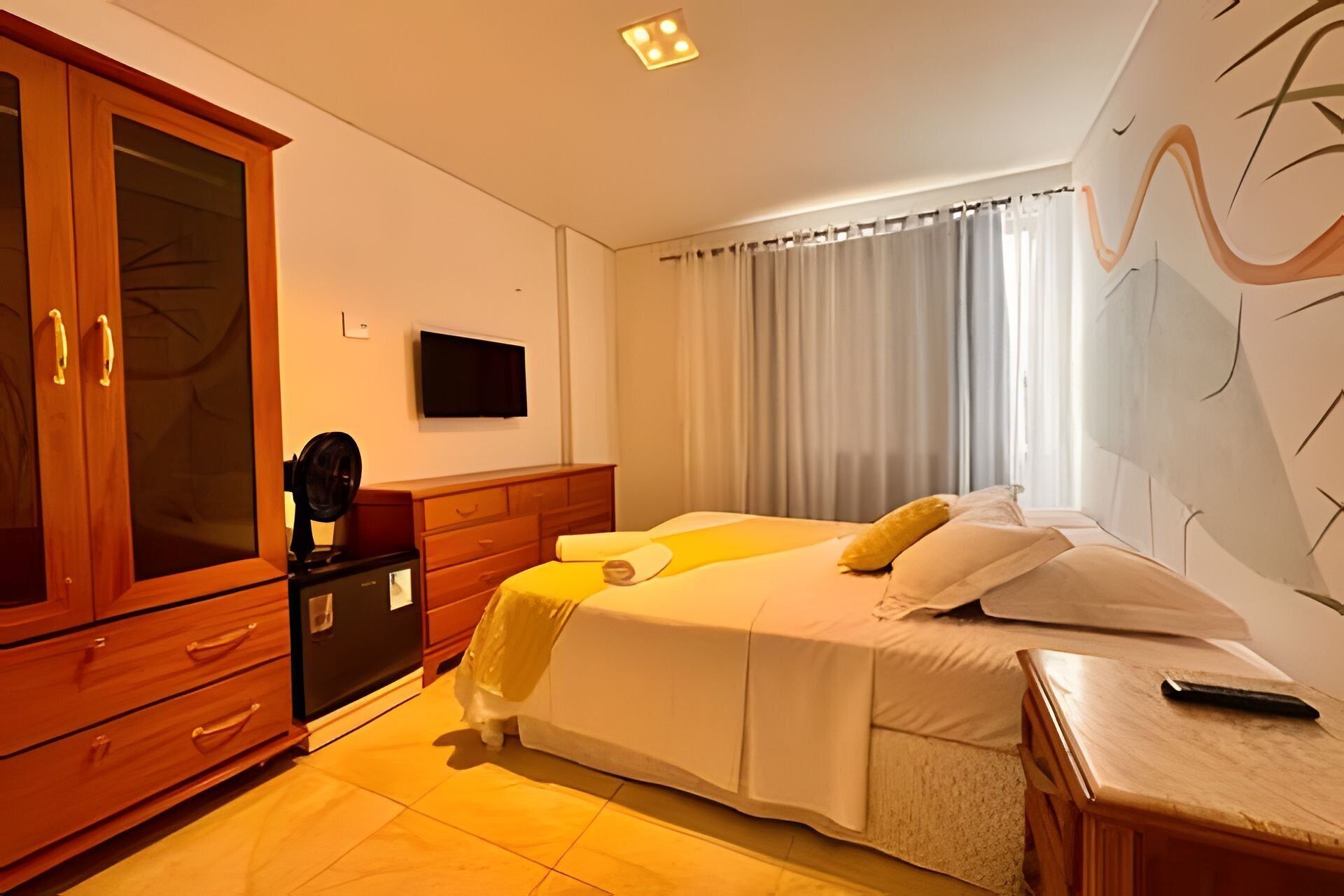 A hotel room with a bed, a wooden wardrobe, a bedside table, and a TV on the wall.