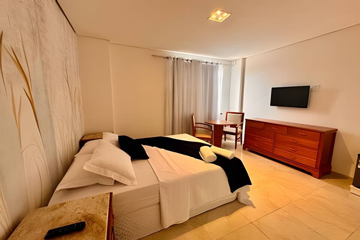 Quarto de hotel com cama, escrivaninha, cadeira, televisão e cómoda de madeira.