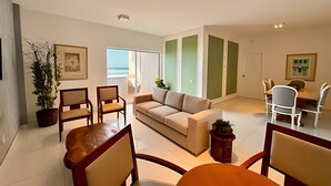 Amplia sala de estar con sofá color beige, sillas de madera, una mesa de comedor con sillas, una planta en maceta y vista al océano a través de una puerta corrediza.