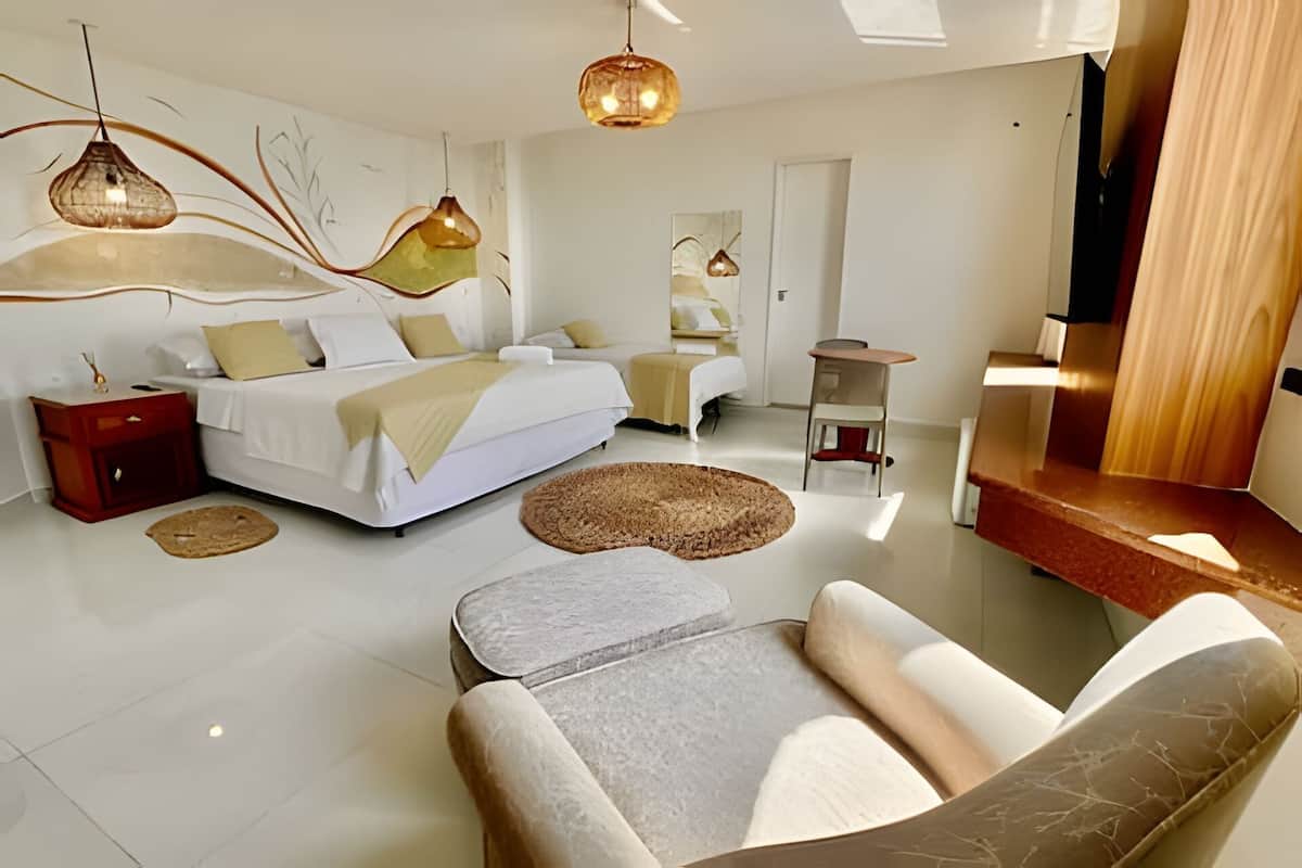 Quarto de hotel moderno com uma cama grande, duas camas menores, uma área de estar com sofá e poltronas, uma escrivaninha e uma televisão.