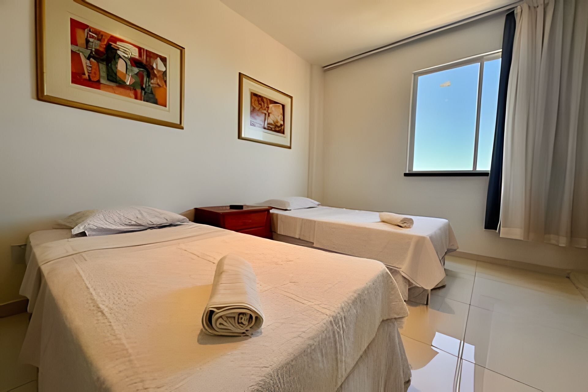 Une chambre d’hôtel avec deux lits, une fenêtre avec des rideaux et deux tableaux encadrés au mur.