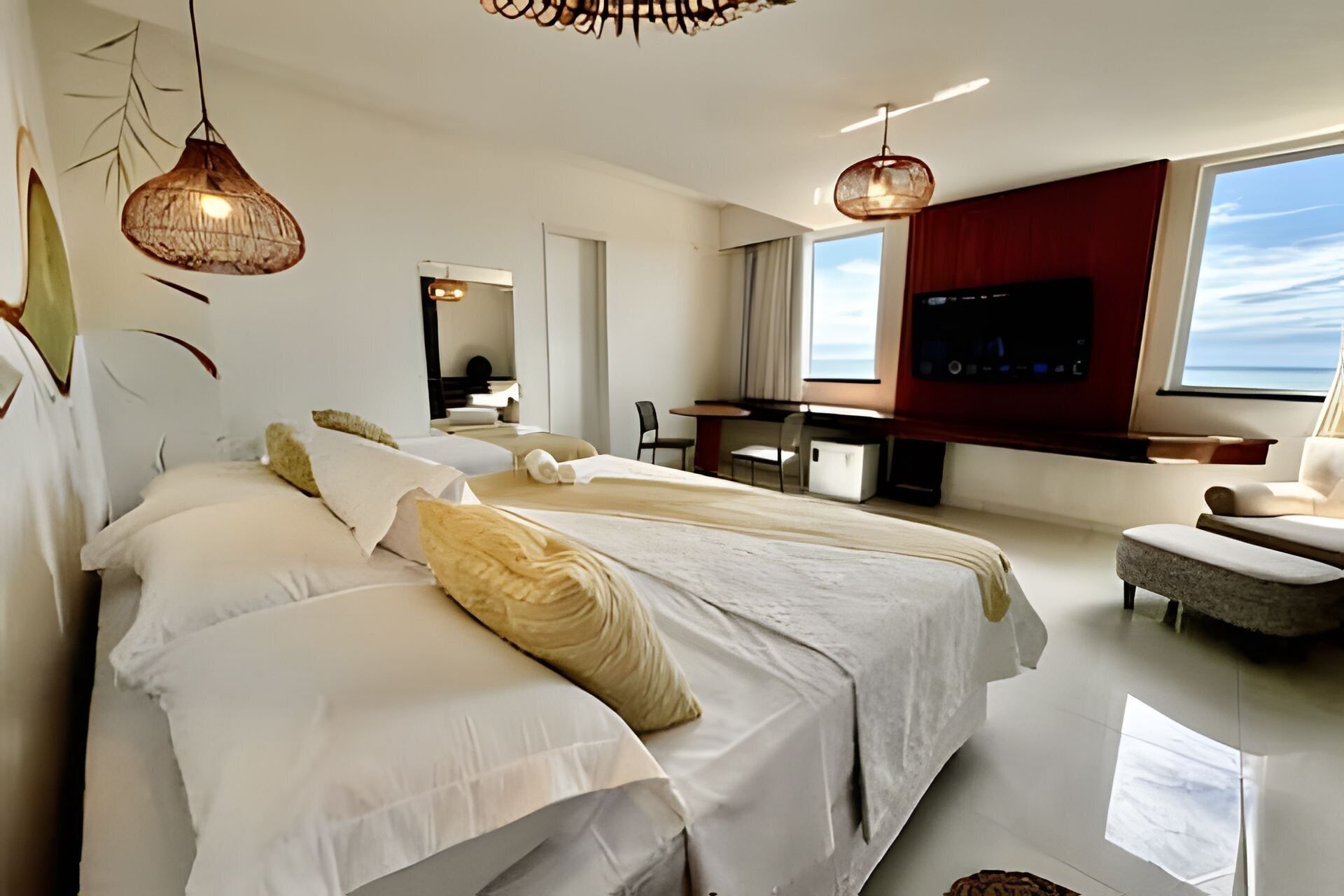 A modern hotel room with two beds, a desk, a TV, and a view of the sea.