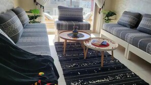 Een woonkamer met een bank, fauteuil en salontafel. Er staan planten en er is uitzicht op gebouwen.