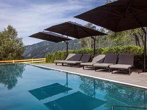 A poolside area with lounge chairs and umbrellas, a wooden fence, and a mountain backdrop. - Ideal Apartment With Shared Sauna (Sankt Martin am Tennengebirge)