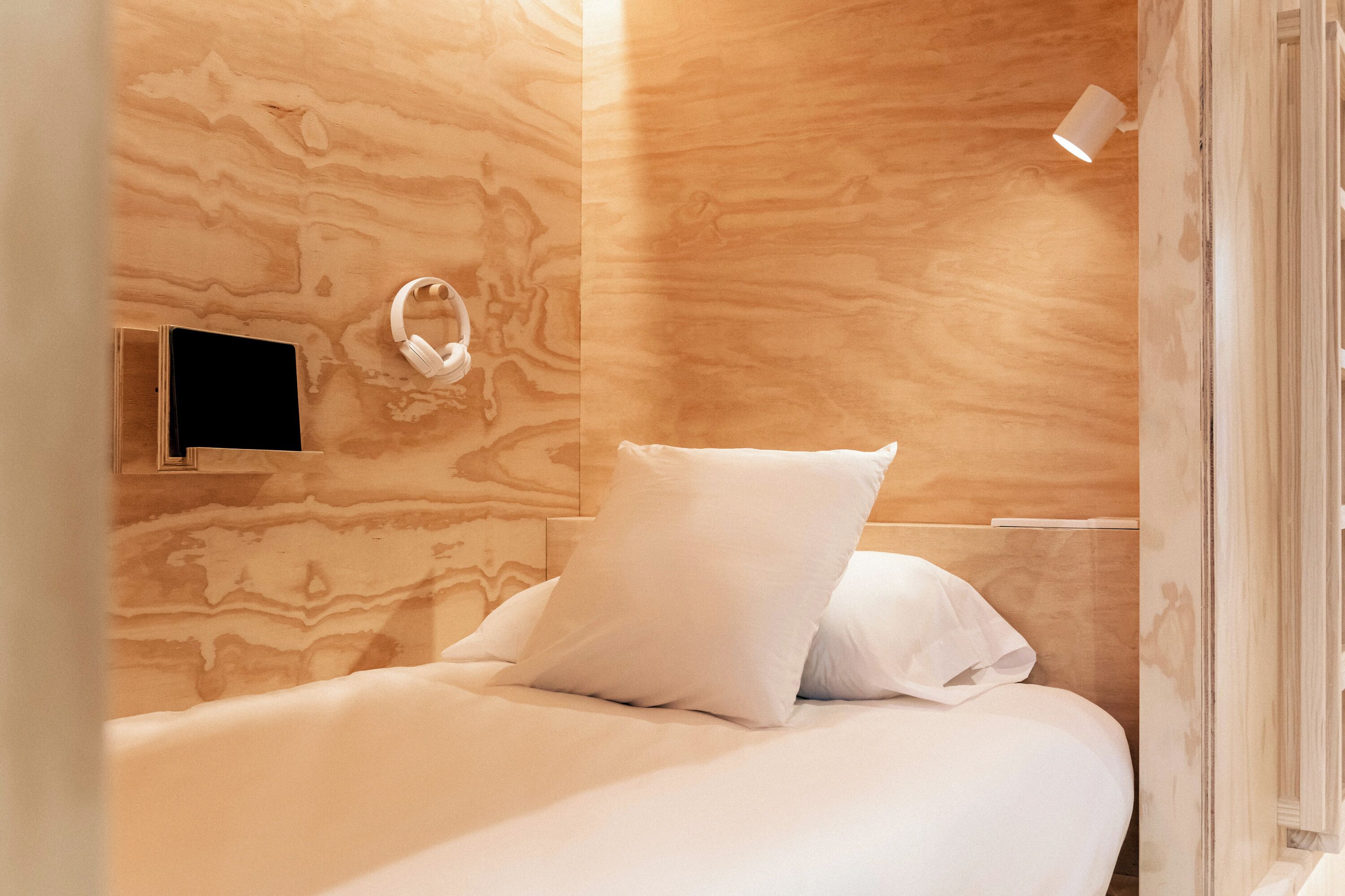 A bedroom with a wooden wall, a bed with white bedding, a white pillow, and a small bedside table with a white lamp.