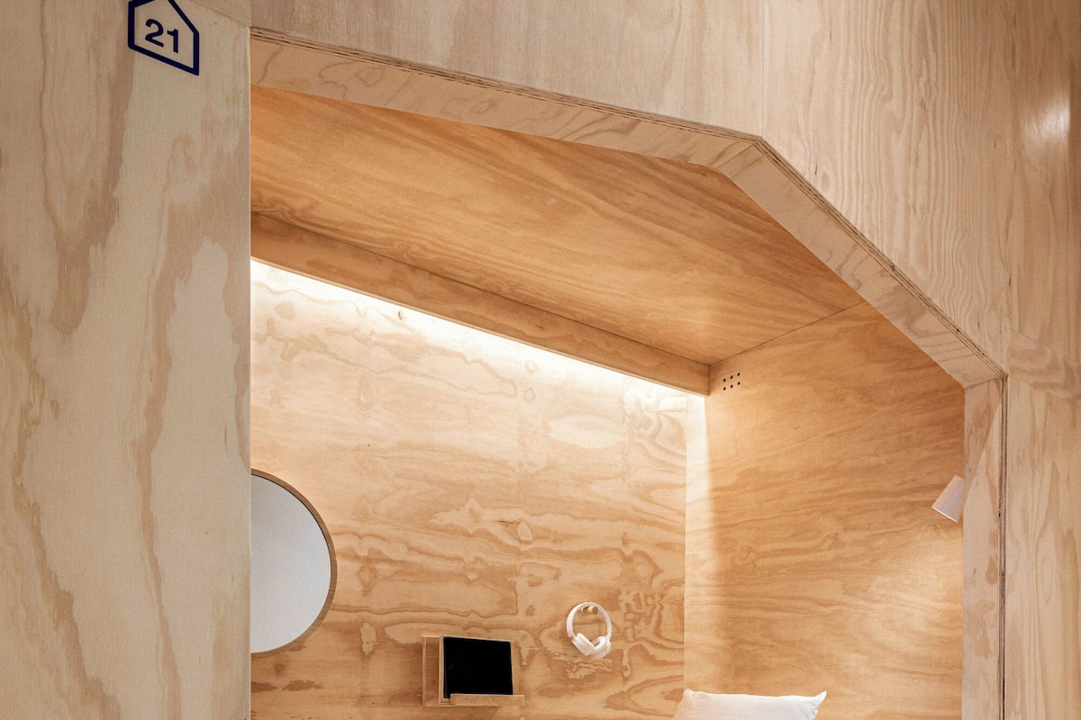 A compact, wooden room with a bed, a circular light fixture, and a small wall-mounted panel.
