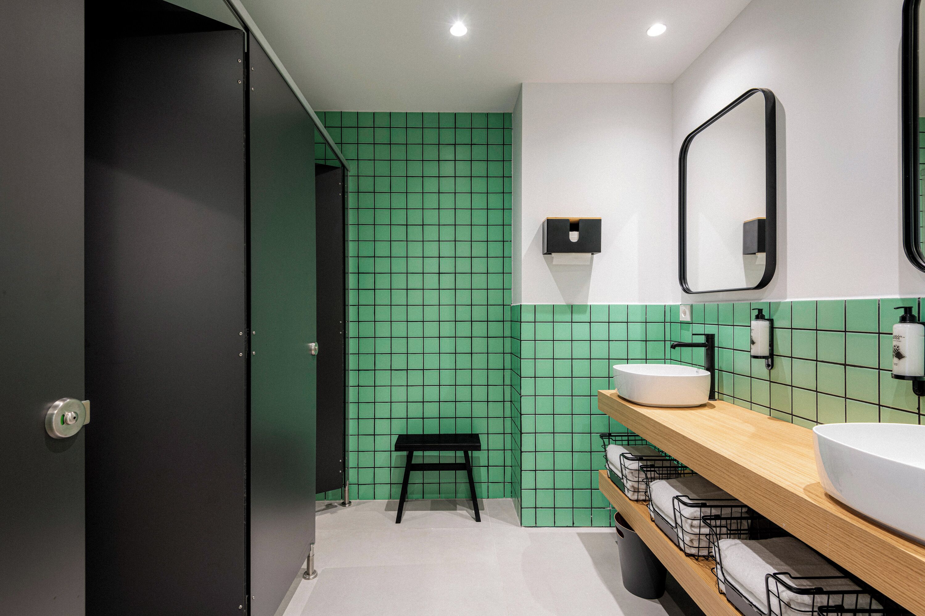 A modern bathroom with a green tiled wall, a wooden vanity with a white sink, and a black bench.