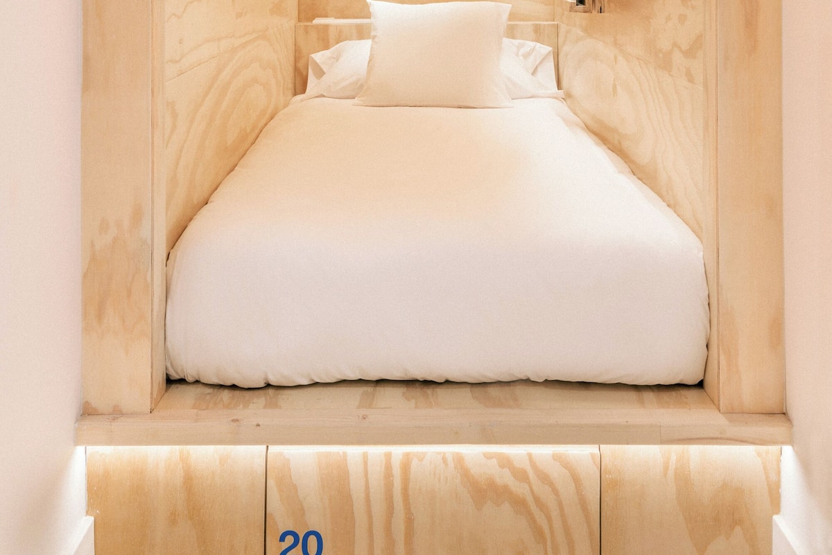 A small, wooden, triangular room with a bed, a mirror, and a small window.