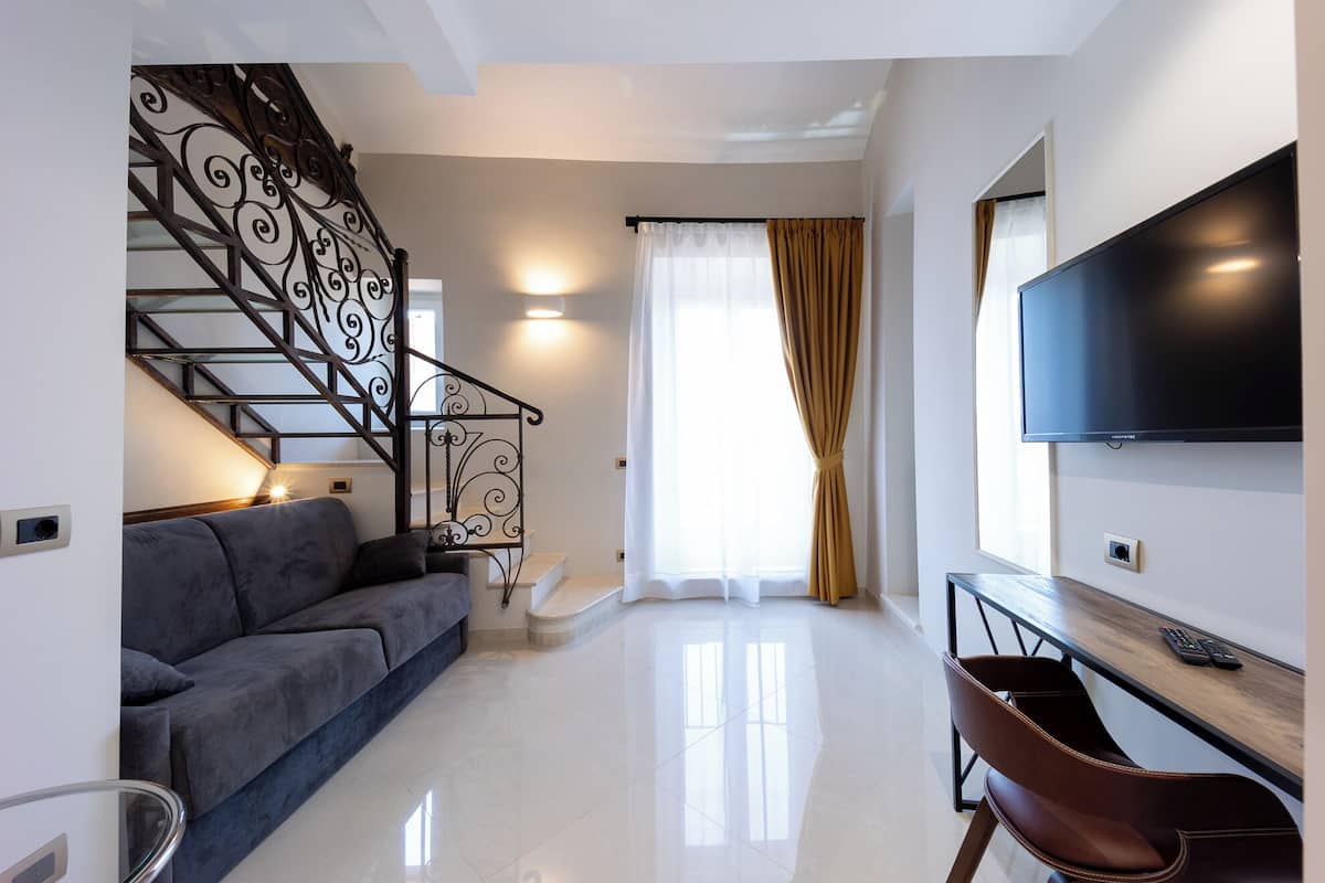 A modern living room with a grey sofa, a wooden desk, a TV, and a staircase with decorative iron railings.