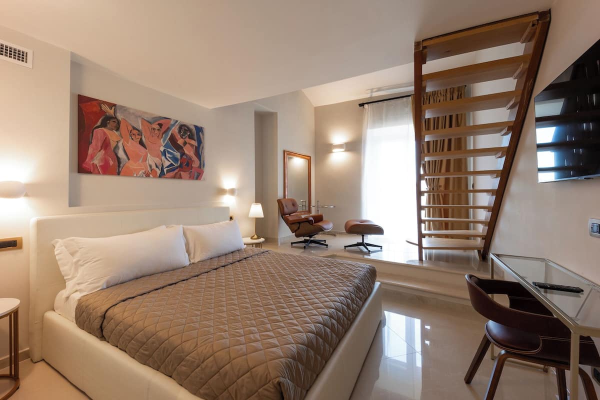 A hotel room with a bed, a desk, a chair, a painting, a mirror, and a TV.
