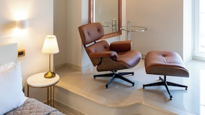 Une chambre d’hôtel moderne avec un lit, un fauteuil en cuir marron, une petite table ronde avec une lampe, une table en verre avec une chaise et un grand miroir.