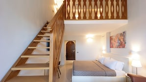Une chambre avec un escalier en bois, un lit avec une literie blanche et grise, un tableau au mur et une lampe sur la table de chevet.