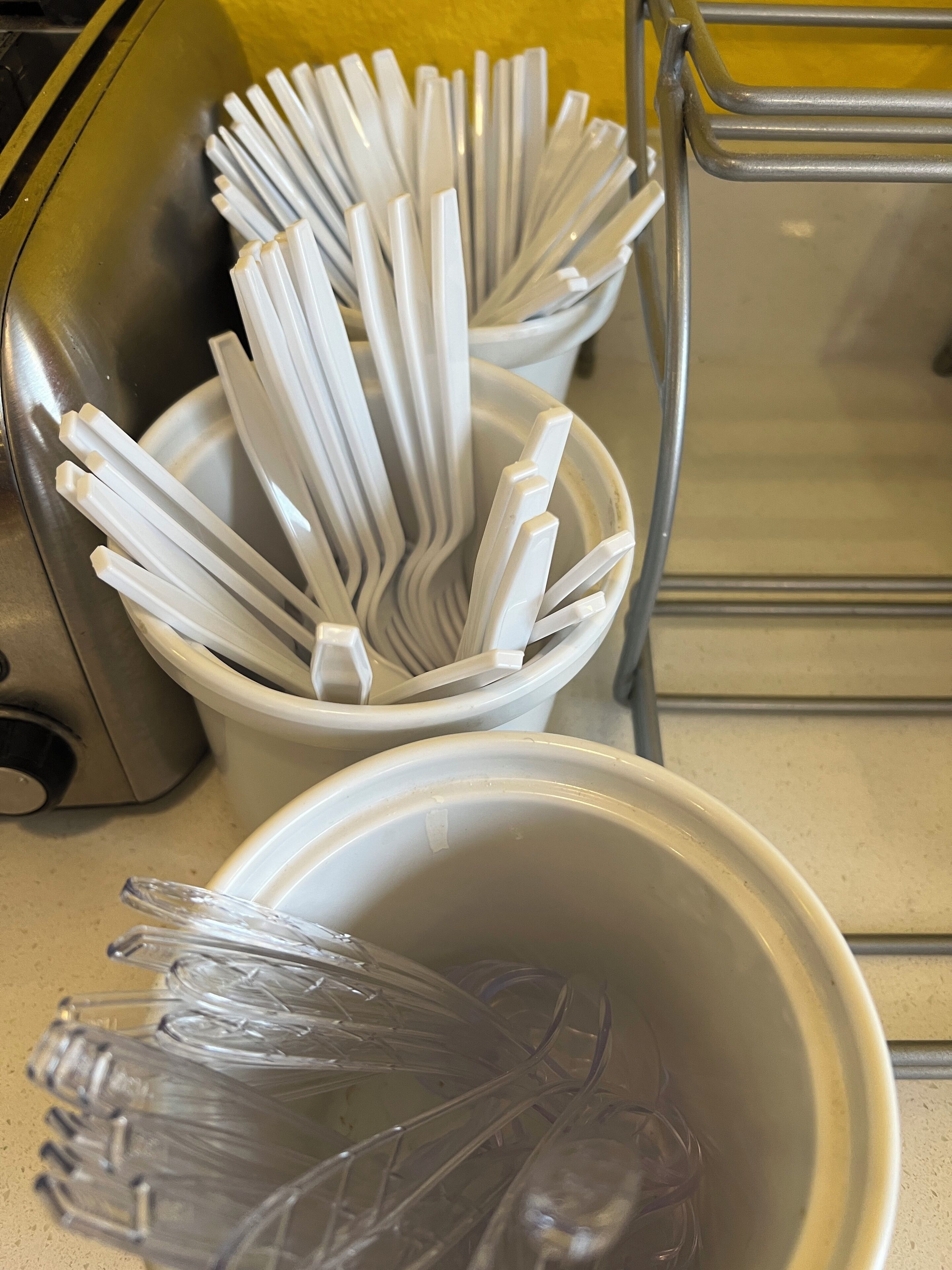 A kitchen area with a stainless steel sink, a white bowl, and a white container holding white plastic utensils.