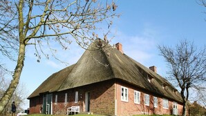 Une maison traditionnelle en briques, avec un toit de chaume, un banc à l’extérieur et une pelouse verdoyante.