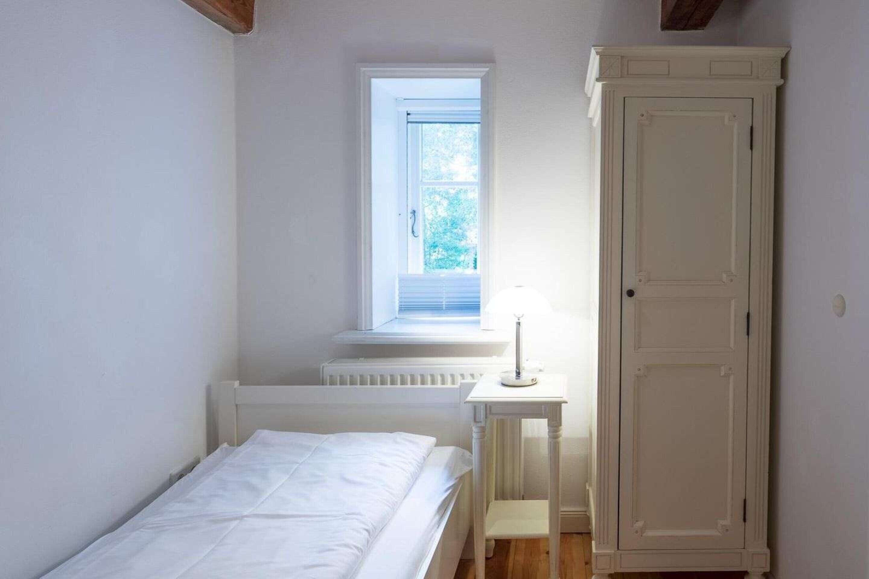 Et lille, hvidt rum med en seng, et vindue, en radiator og et hvidt skab.
