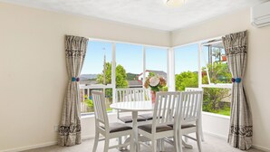 Una habitación con una mesa y sillas blancas para comer, ventanas amplias con cortinas y vistas al exterior con zonas verdes y casas.