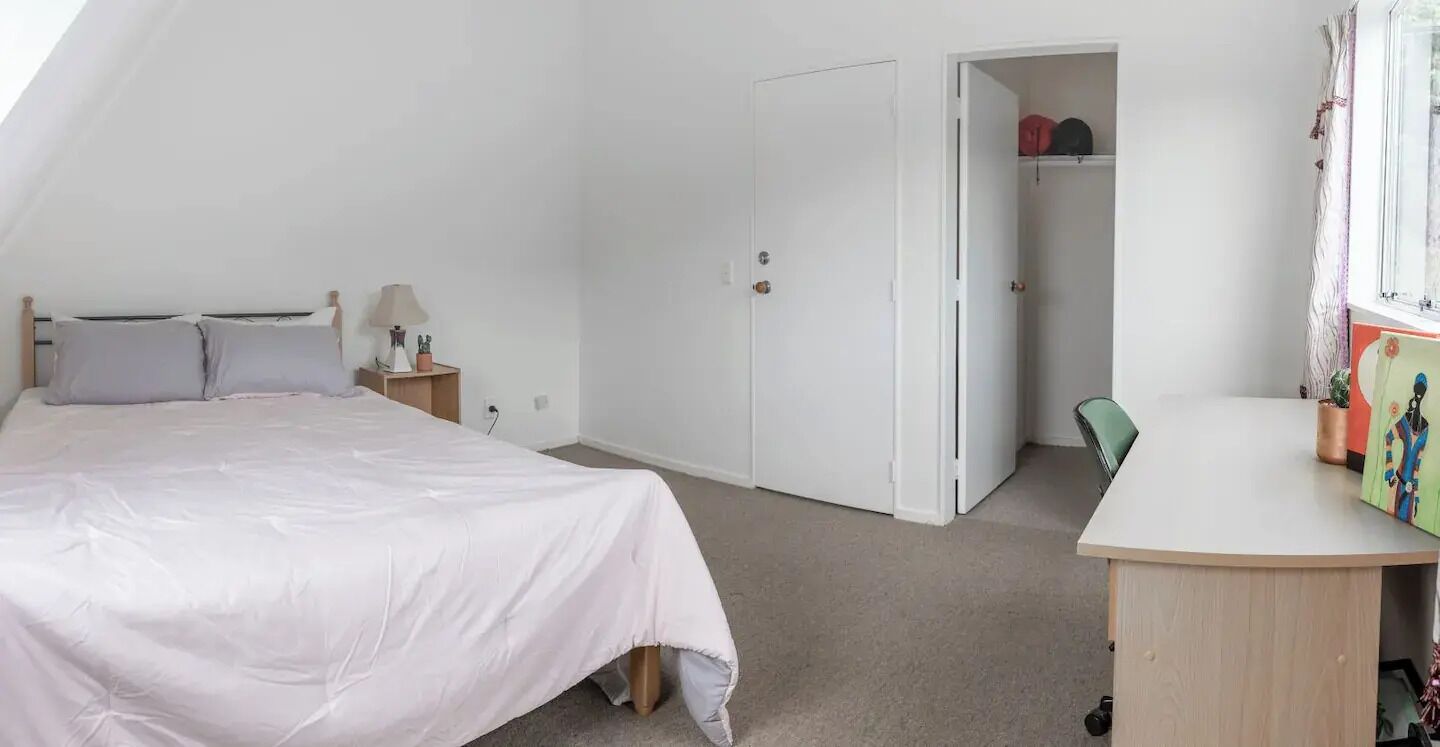 Un dormitorio con cama, mesita de noche, lámpara y escritorio con silla. Hay un armario y una puerta que da a otra habitación.