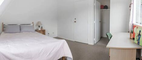 Un dormitorio con cama, mesita de noche, lámpara y escritorio con silla. Hay un armario y una puerta que da a otra habitación.