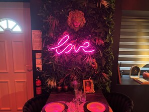 A room with a neon sign, a lion head decoration, a framed picture, and a table with glasses and a plate. - La Sainteny Touch - Erotic Room (Terre-et-Marais)