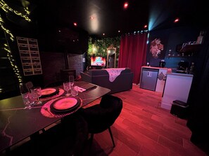 A dimly lit room with a dining table set for two, a sofa, and a kitchen area with a refrigerator and a coffee machine.