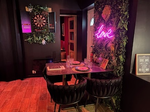 A dark-themed room with a dartboard, neon 'love' sign, and a framed quote on the wall.
